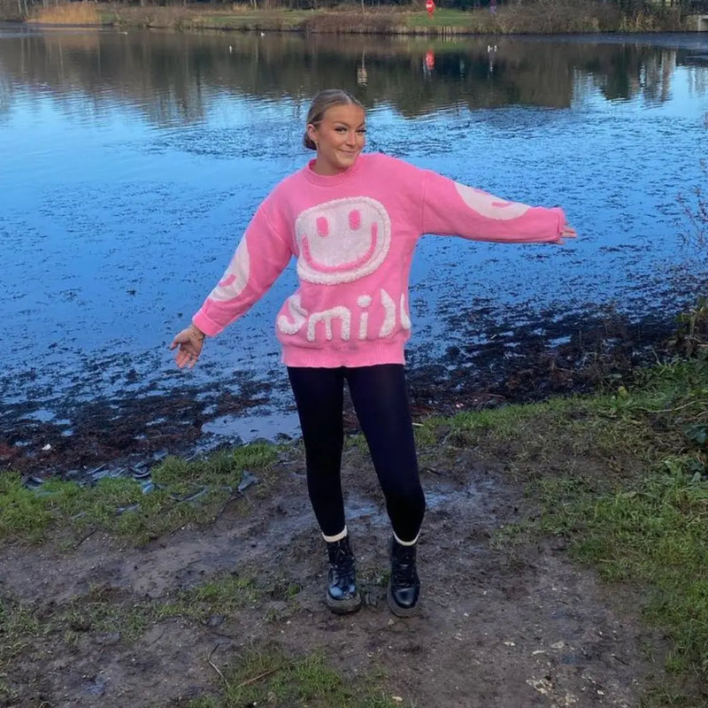 A customer by a lake wearing a pink knitted sweater, arms out